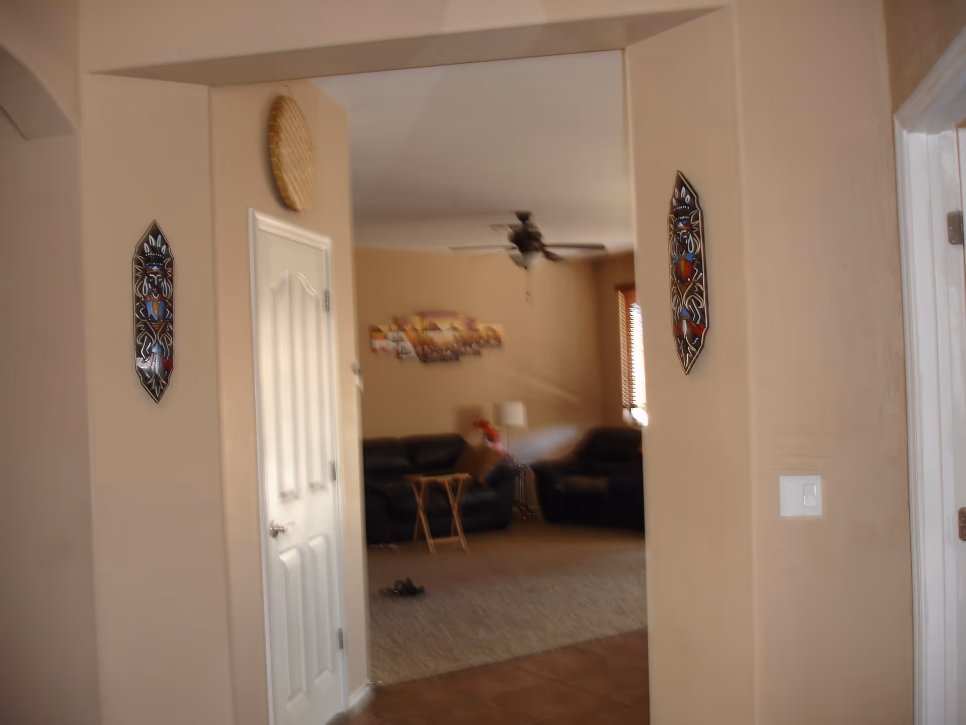 View through a hallway into a living room with beige walls, two black leather sofas, a small wooden folding table, a ceiling fan, and wall decorations including two tribal masks and a woven basket.