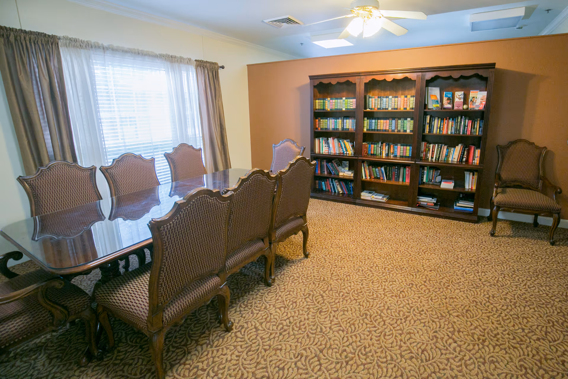 A room with a long wooden table surrounded by eight upholstered chairs. There is a large window with sheer and heavy curtains on one wall, and a wooden bookshelf filled with books and pamphlets against another wall. The room has patterned carpet and a ceiling fan with lights.
