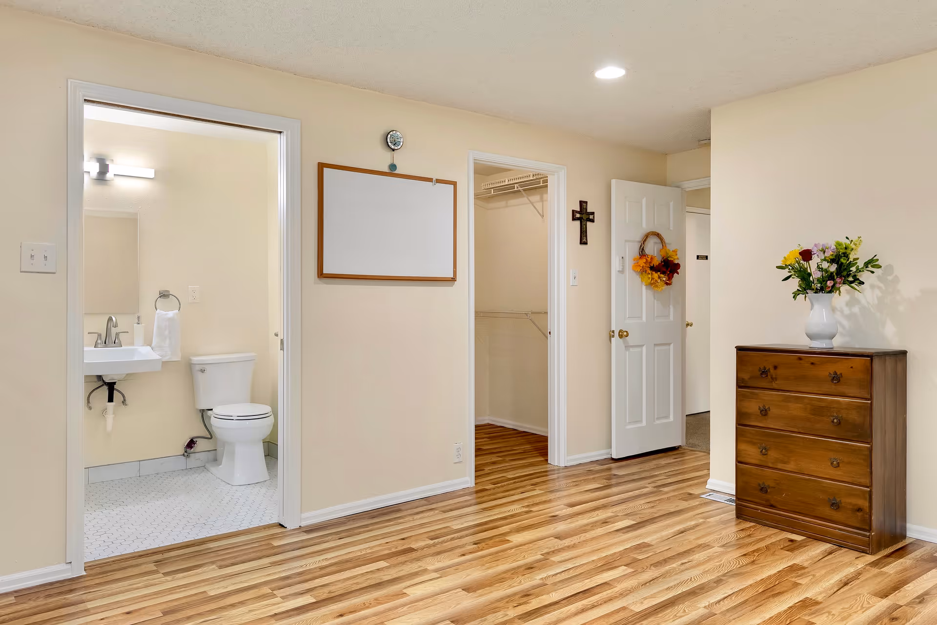 Interior view of a room showing a bathroom doorway, an open closet, a wooden dresser with a vase of flowers, and hardwood flooring.