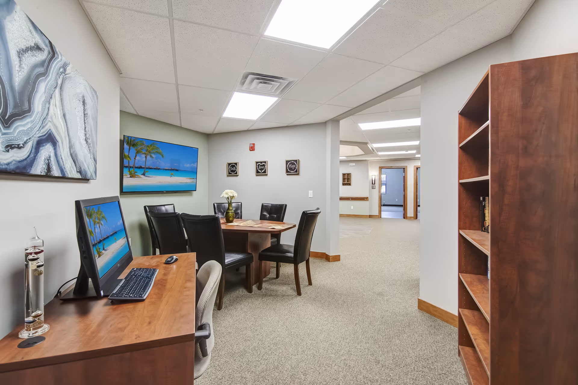 Interior common area with a computer desk, a round table surrounded by chairs, wall art, and a wooden bookshelf.