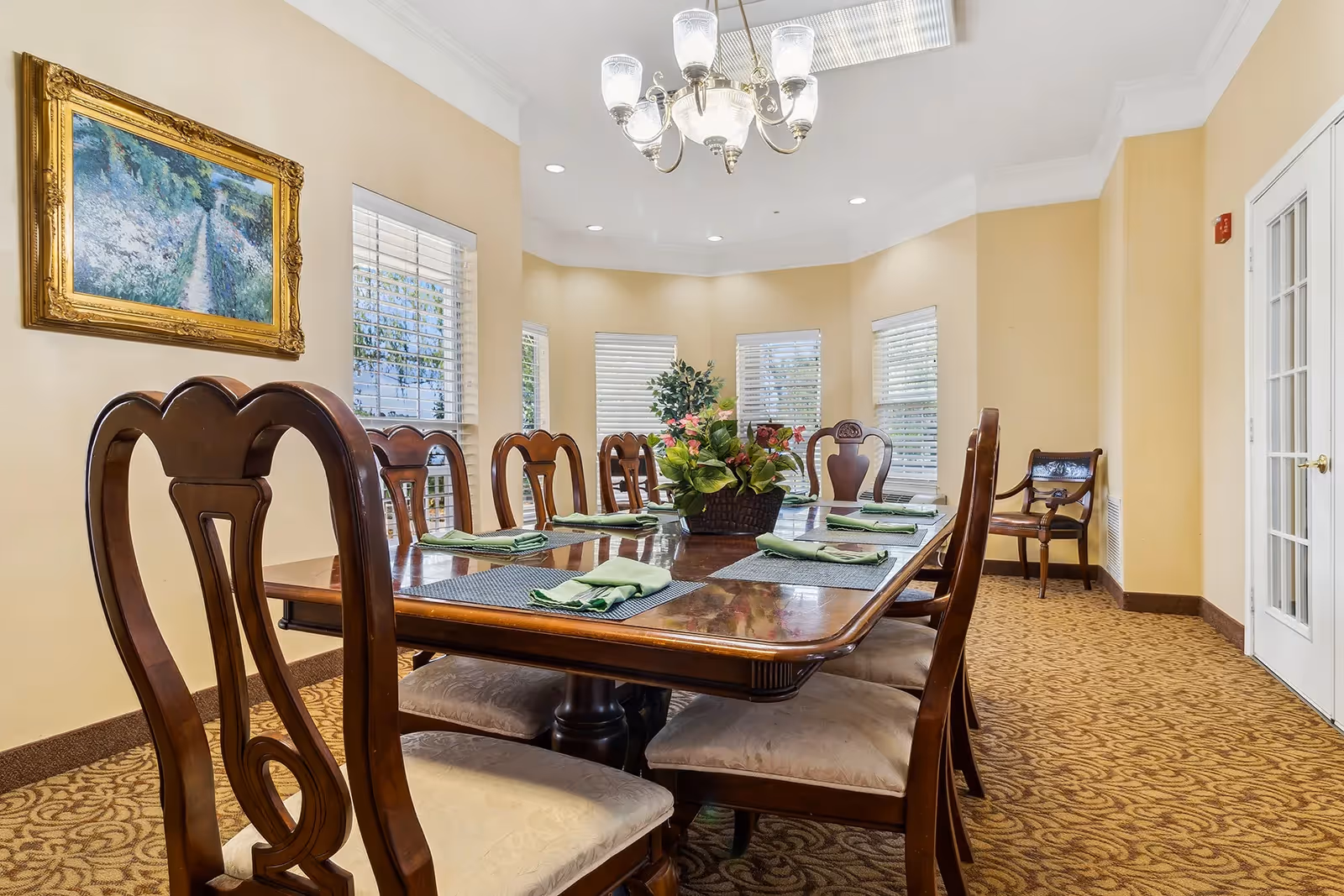 Formal dining room with a long wooden table and chairs, place settings and a floral centerpiece under a chandelier beside windows.
