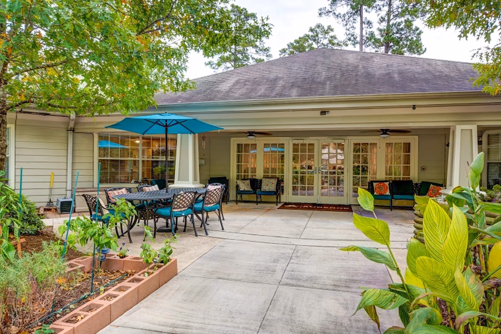Outdoor patio area at The Auberge at The Woodlands featuring a table with six chairs under a blue umbrella, surrounded by greenery and plants, with a building entrance in the background having glass double doors and windows.