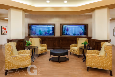 A cozy seating area with four yellow patterned armchairs arranged around a round black ottoman. Behind the chairs, there are two large built-in aquariums set into a wooden panel wall. The space has warm lighting and a wood floor.