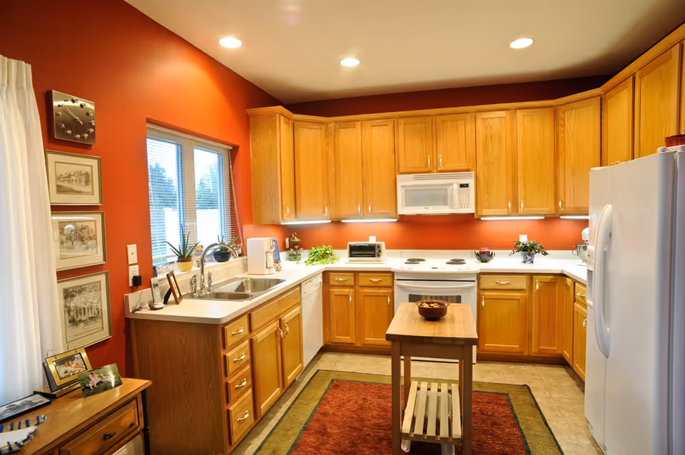 A well-lit kitchen with wooden cabinets, white countertops, and white appliances including a refrigerator, stove, microwave, and dishwasher. There is a small wooden island in the center with a bowl on top. The walls are painted a warm red color, and there is a window above the sink with blinds. Various plants and framed pictures decorate the space.