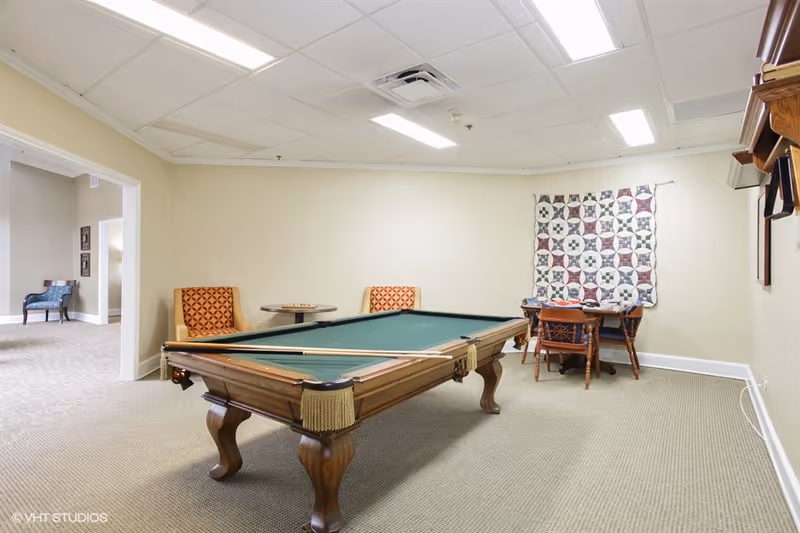 A recreational room with a pool table in the center, two orange patterned chairs and a small round table against the wall, a wooden table with four chairs, and a quilt hanging on the wall. The room has beige walls and carpeted floor, with fluorescent ceiling lights.