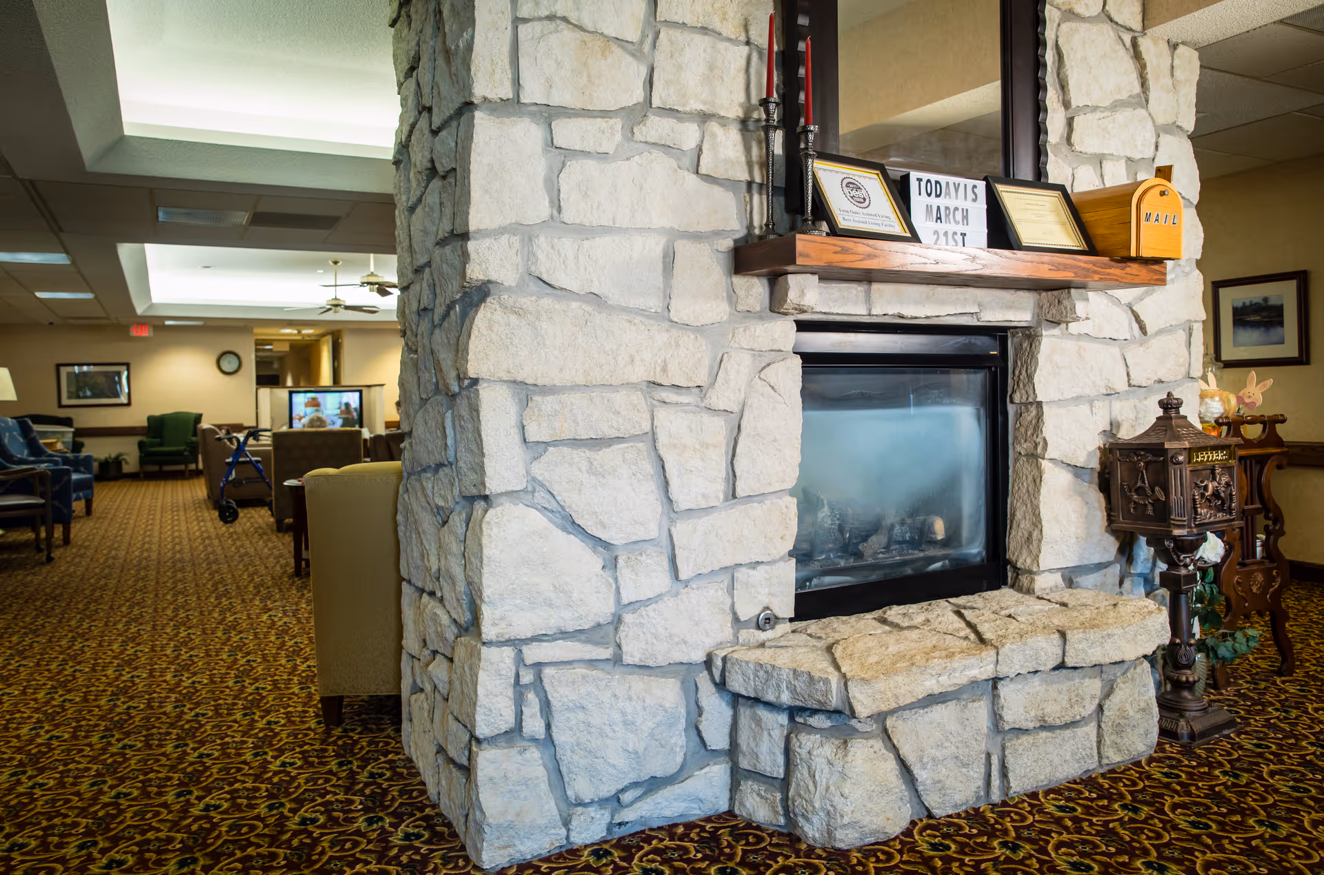 A stone fireplace in a senior living facility lounge with chairs and patterned carpet visible in the background.