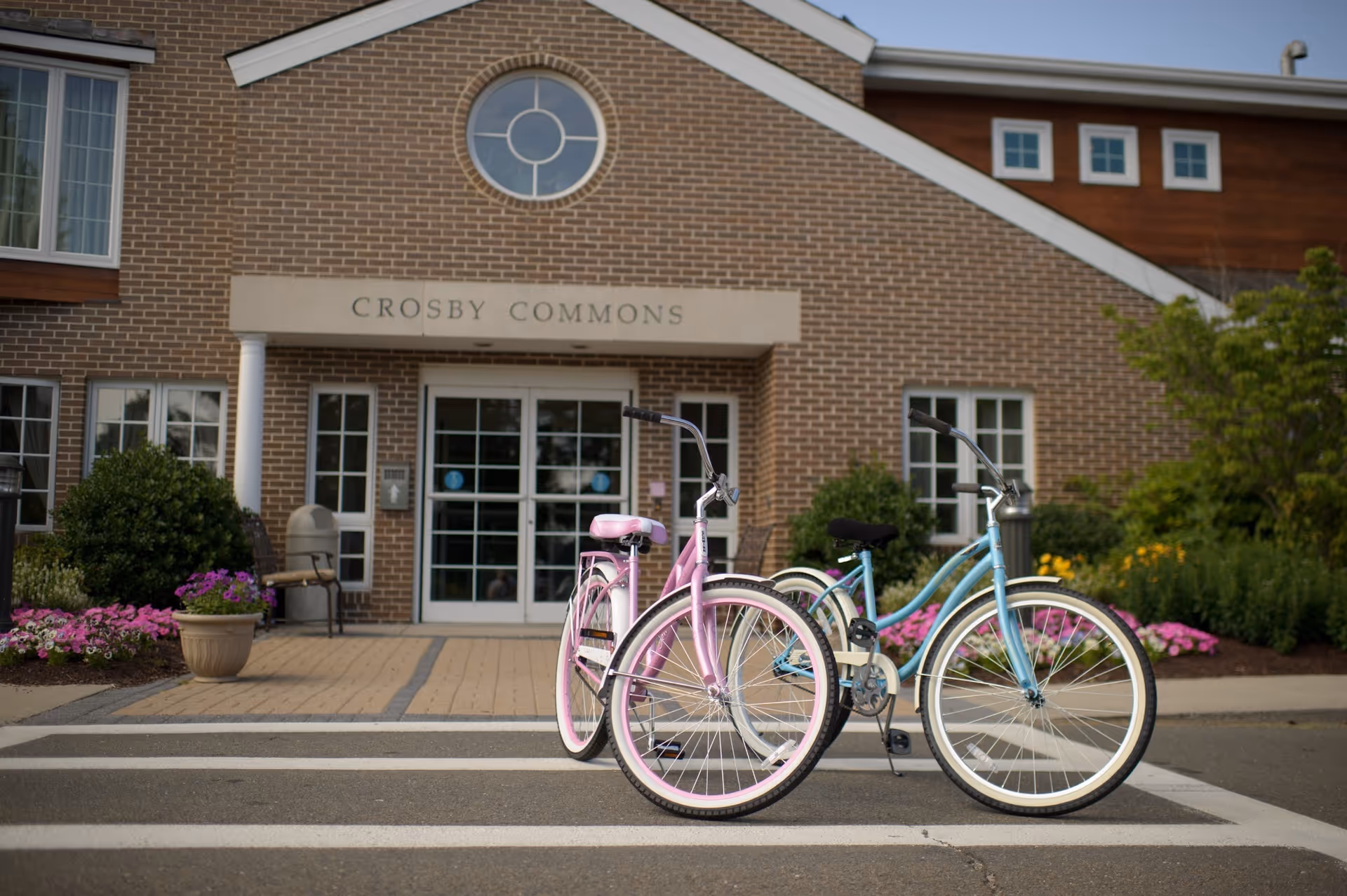 Two bicycles, one pink and one blue, parked on a crosswalk in front of the brick building entrance of Crosby Commons, with flower beds and greenery on either side.
