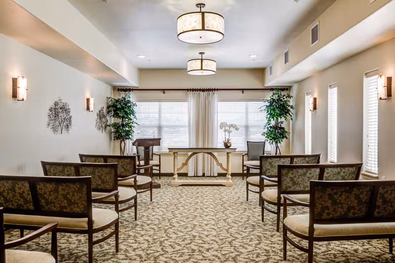 A well-lit room with patterned carpet and floral upholstered chairs arranged in rows facing a white decorative table and a wooden podium near large windows with white curtains. The walls are light-colored with metal wall art and sconces, and there are two tall green plants on either side of the table. Two ceiling light fixtures provide additional lighting.