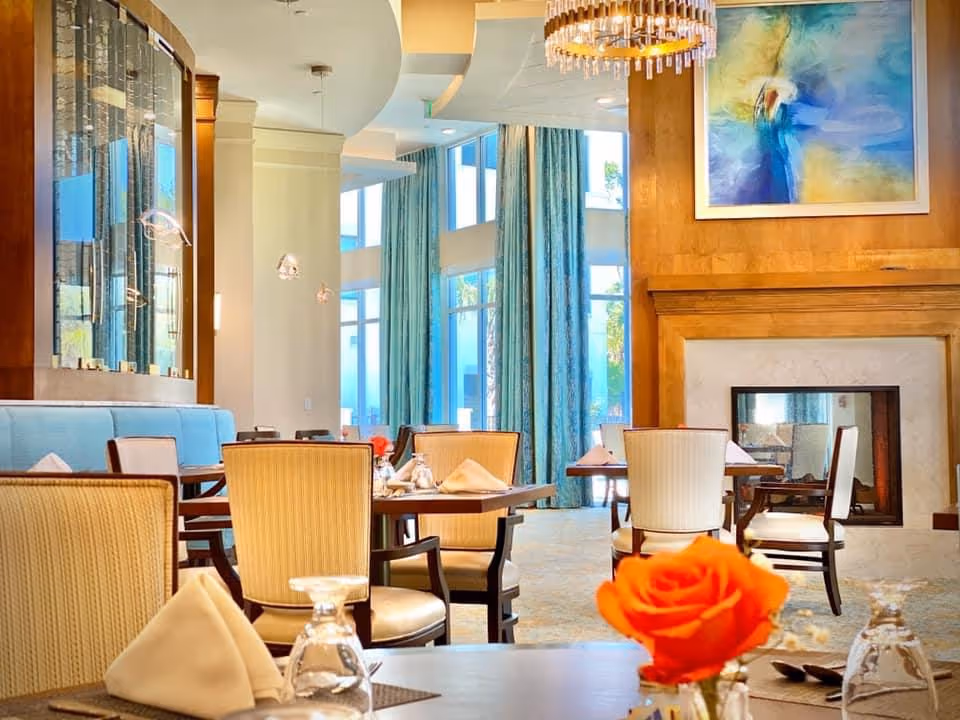 Bright dining room with set tables and chairs, a fireplace and large windows with blue curtains, an orange rose centerpiece in the foreground.