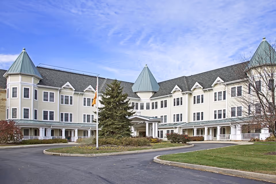 Exterior view of a large senior living facility building with three stories, light-colored siding, green peaked roofs, and multiple windows. There is a circular driveway with a flagpole and landscaped greenery in front of the entrance.