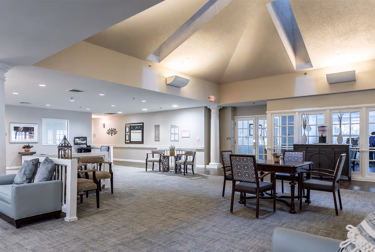 A spacious and well-lit common area in a senior living facility with multiple seating arrangements including armchairs and tables with chairs. The room features a high ceiling with skylights, neutral-colored walls, carpeted floor, decorative columns, and framed artwork on the walls. There are glass doors and windows leading to another room in the background.