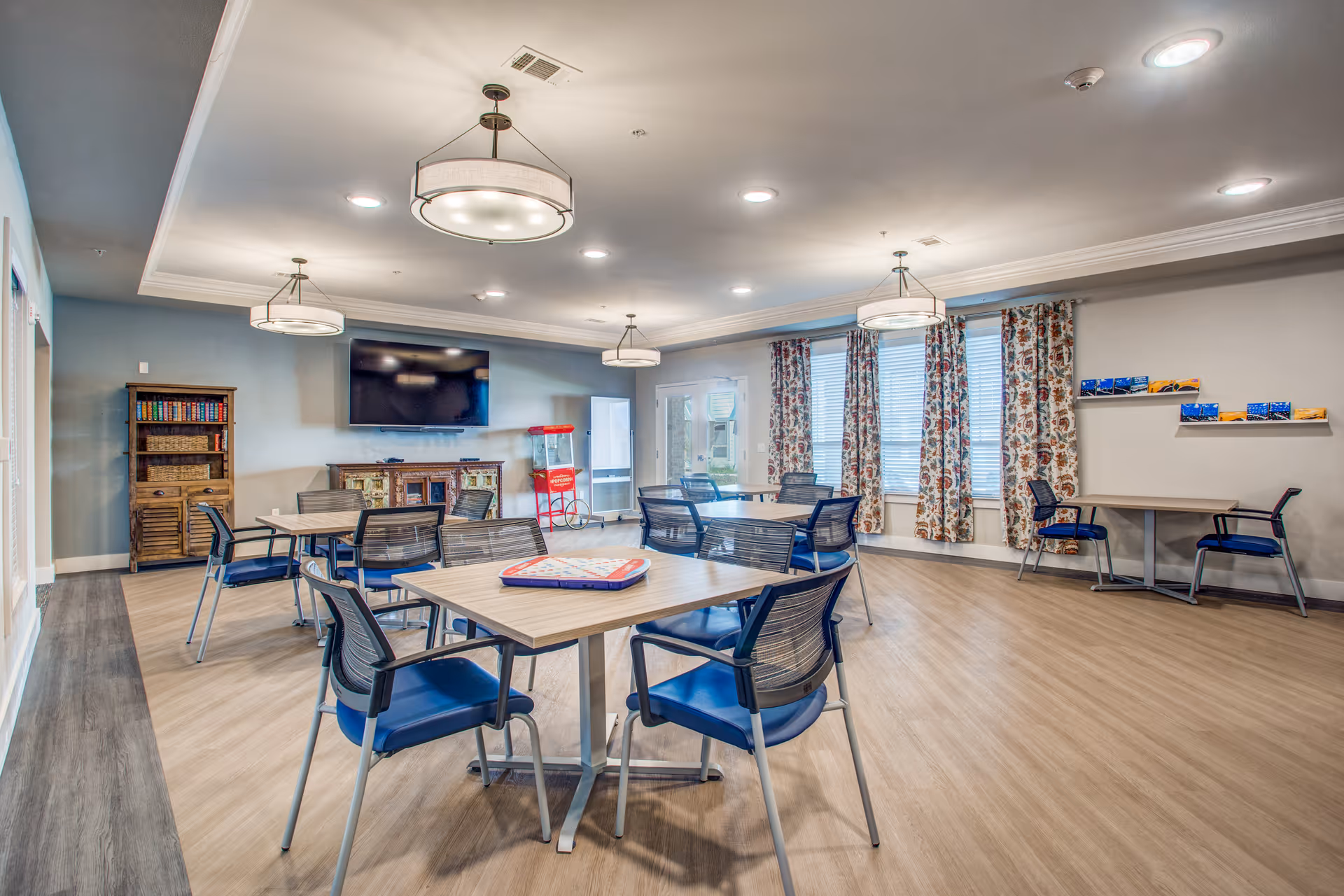 A spacious and well-lit common room with multiple tables and chairs arranged for group activities or dining. The room features a large flat-screen TV mounted on the wall, a popcorn machine, a wooden cabinet with books, and windows with floral curtains allowing natural light to enter. The ceiling has modern light fixtures and recessed lighting.
