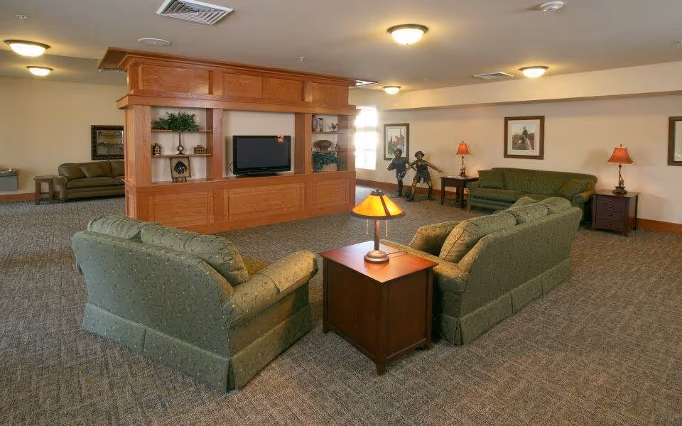 Spacious lounge area with green sofas arranged around a wooden entertainment center holding a TV, side tables and lamps.