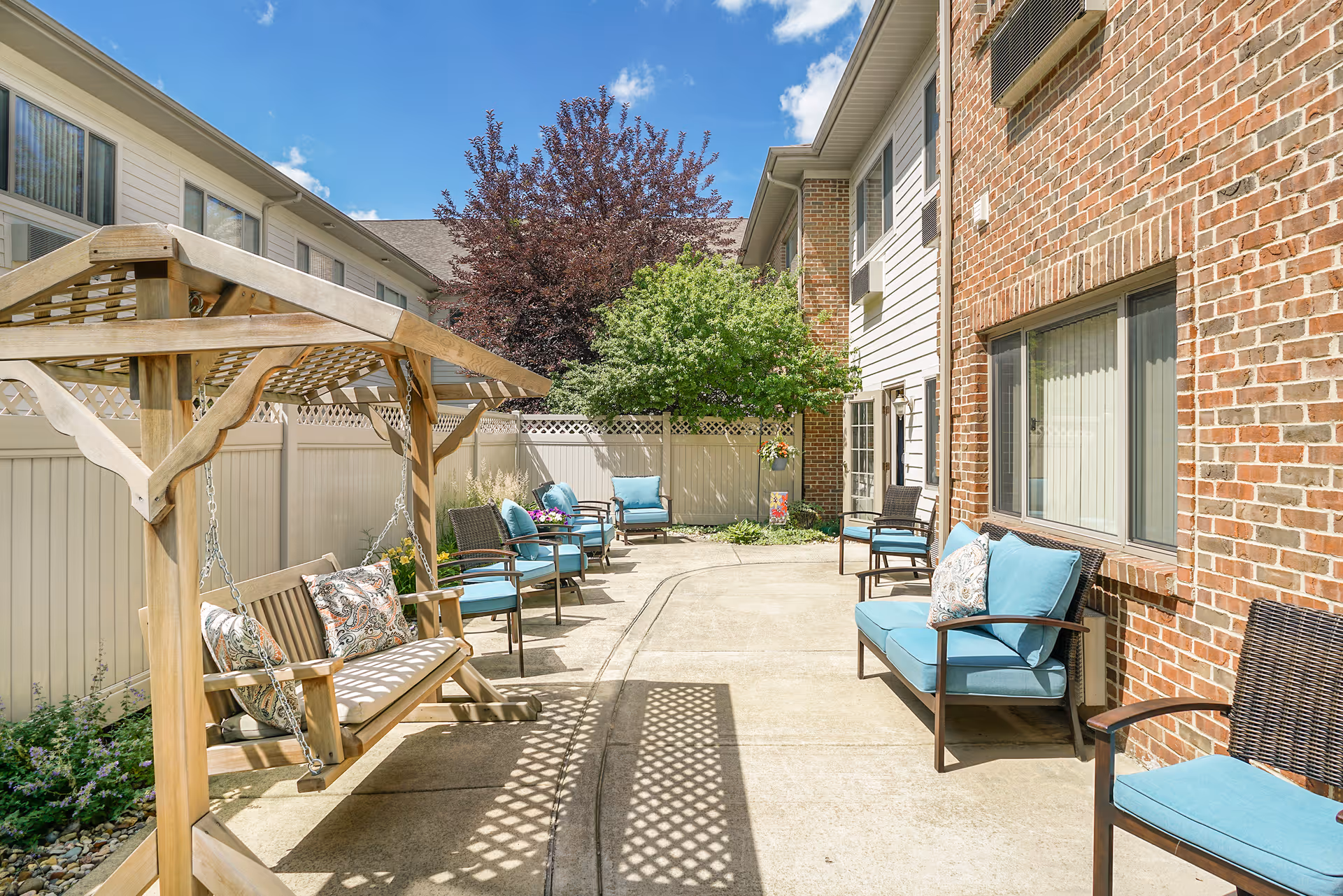 Outdoor patio area with cushioned chairs and a wooden swing bench under a pergola, surrounded by a white fence and brick building walls, with trees and blue sky in the background.