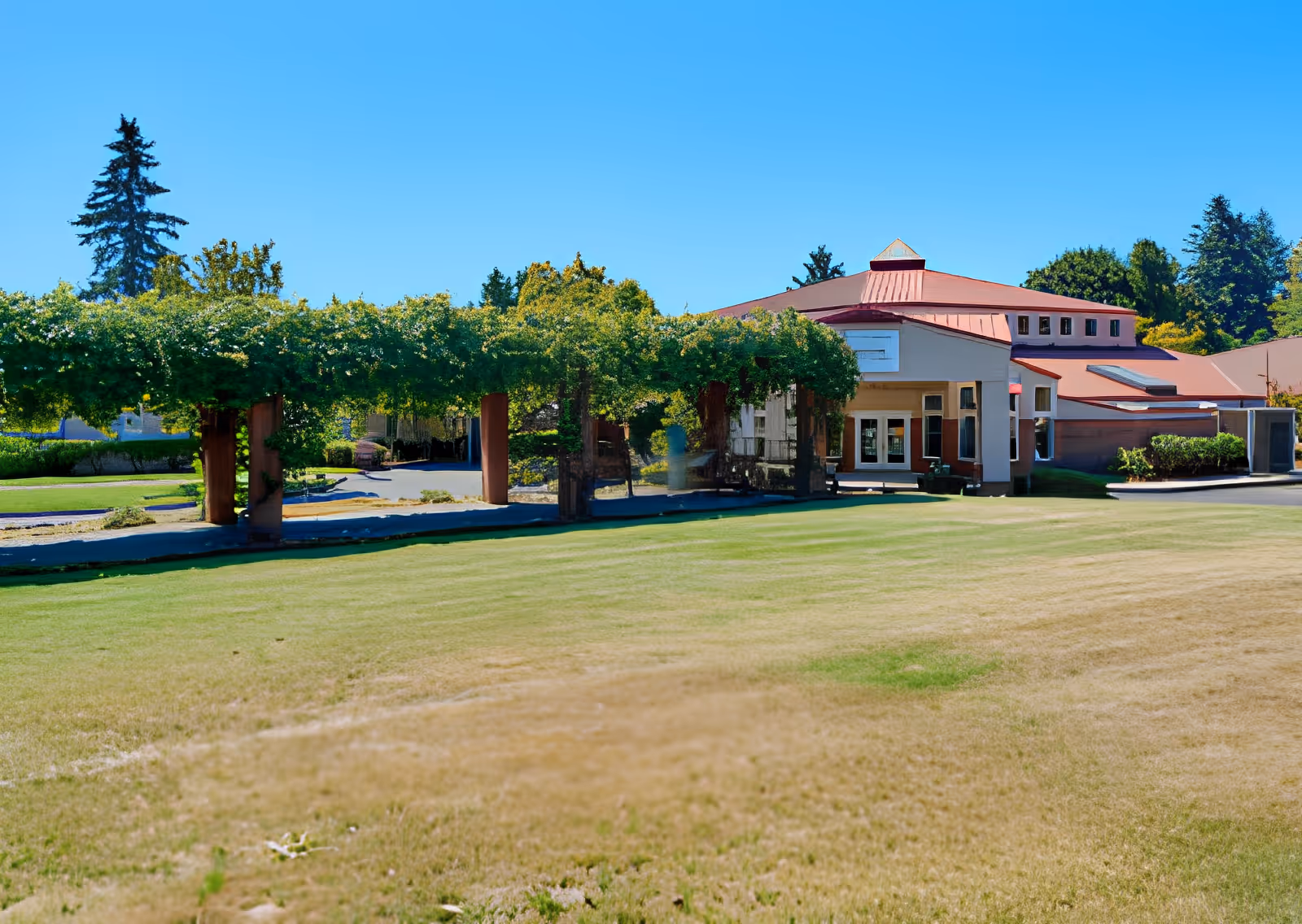 Exterior view of Elliott Residence showing a large green lawn in the foreground, a pergola covered with greenery, and a building with a red roof and multiple windows under a clear blue sky.