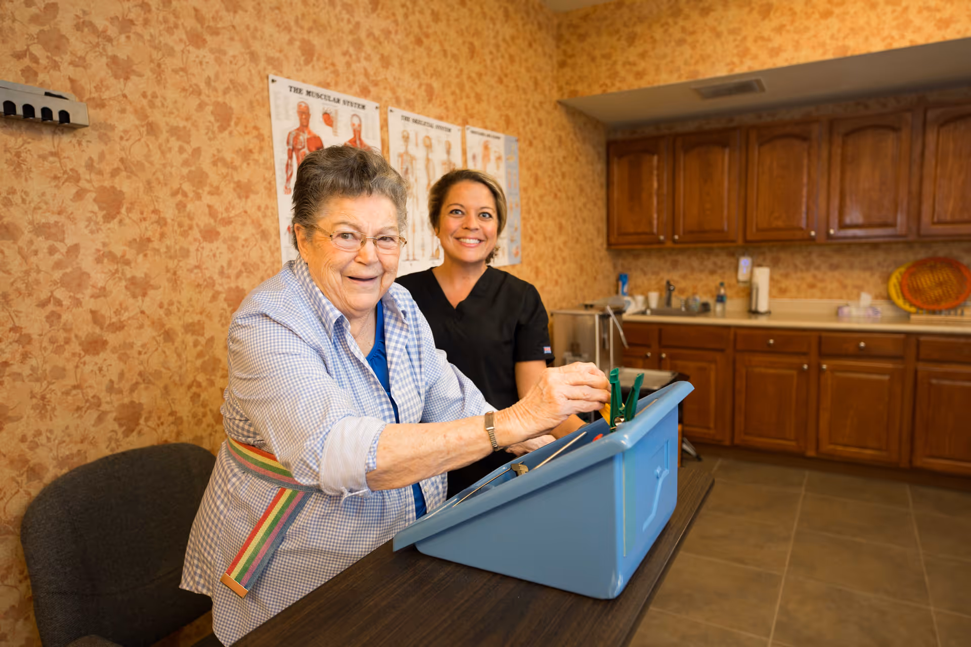 An elderly woman and a healthcare worker smiling together in a room with wooden cabinets and anatomical posters on the wall. The elderly woman is engaging with a blue activity box on the table.