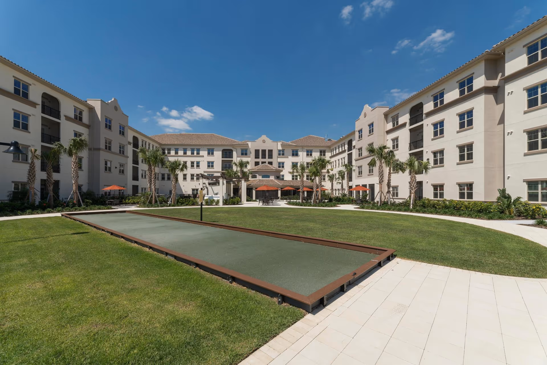 Outdoor courtyard area of a senior living facility with a bocce ball court in the center, surrounded by well-maintained green grass, palm trees, and a multi-story beige building with balconies and windows under a clear blue sky.