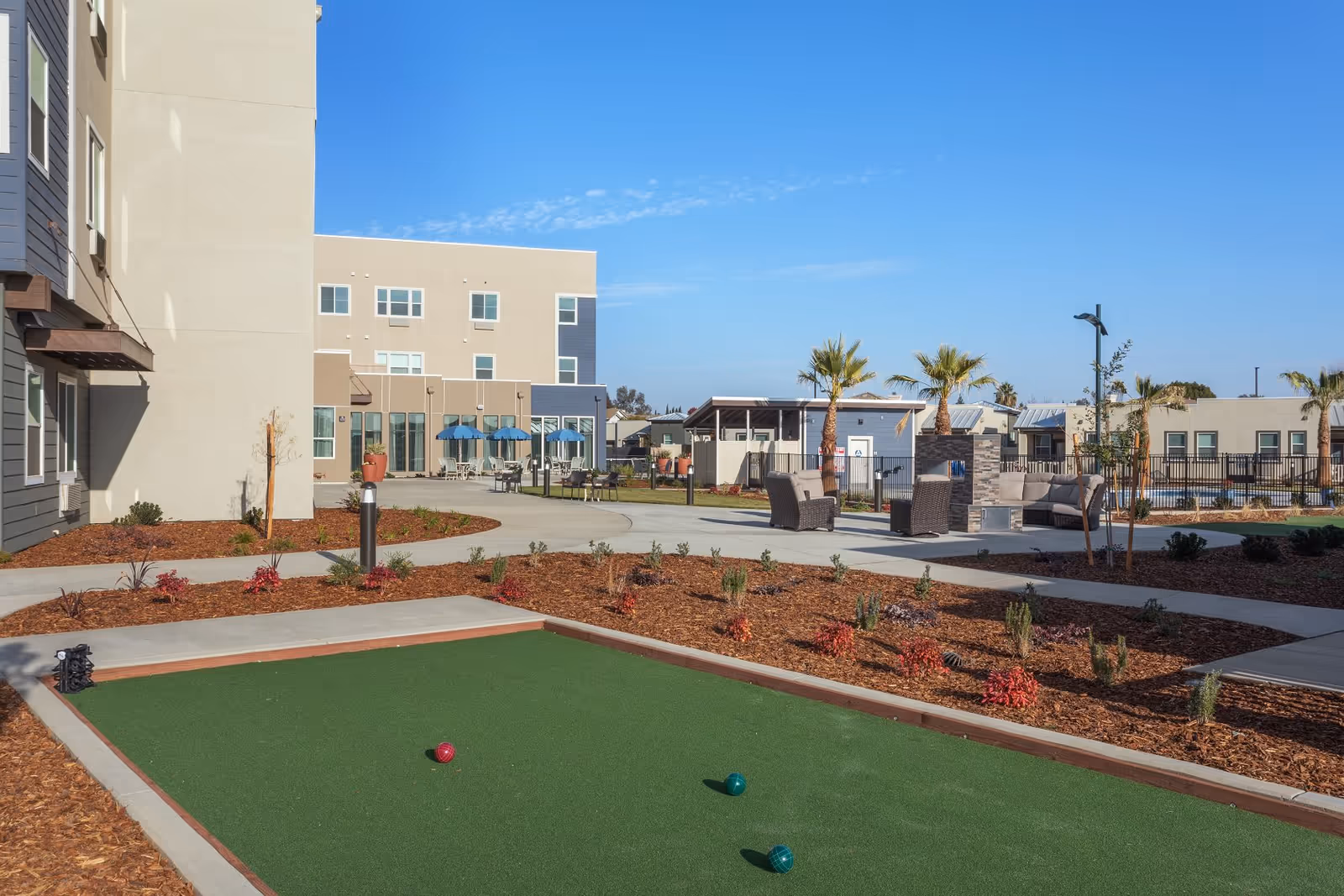 Courtyard at a senior living community showing a small bocce/putting green, planted beds, outdoor seating, palm trees and a multi-story building under a clear blue sky.