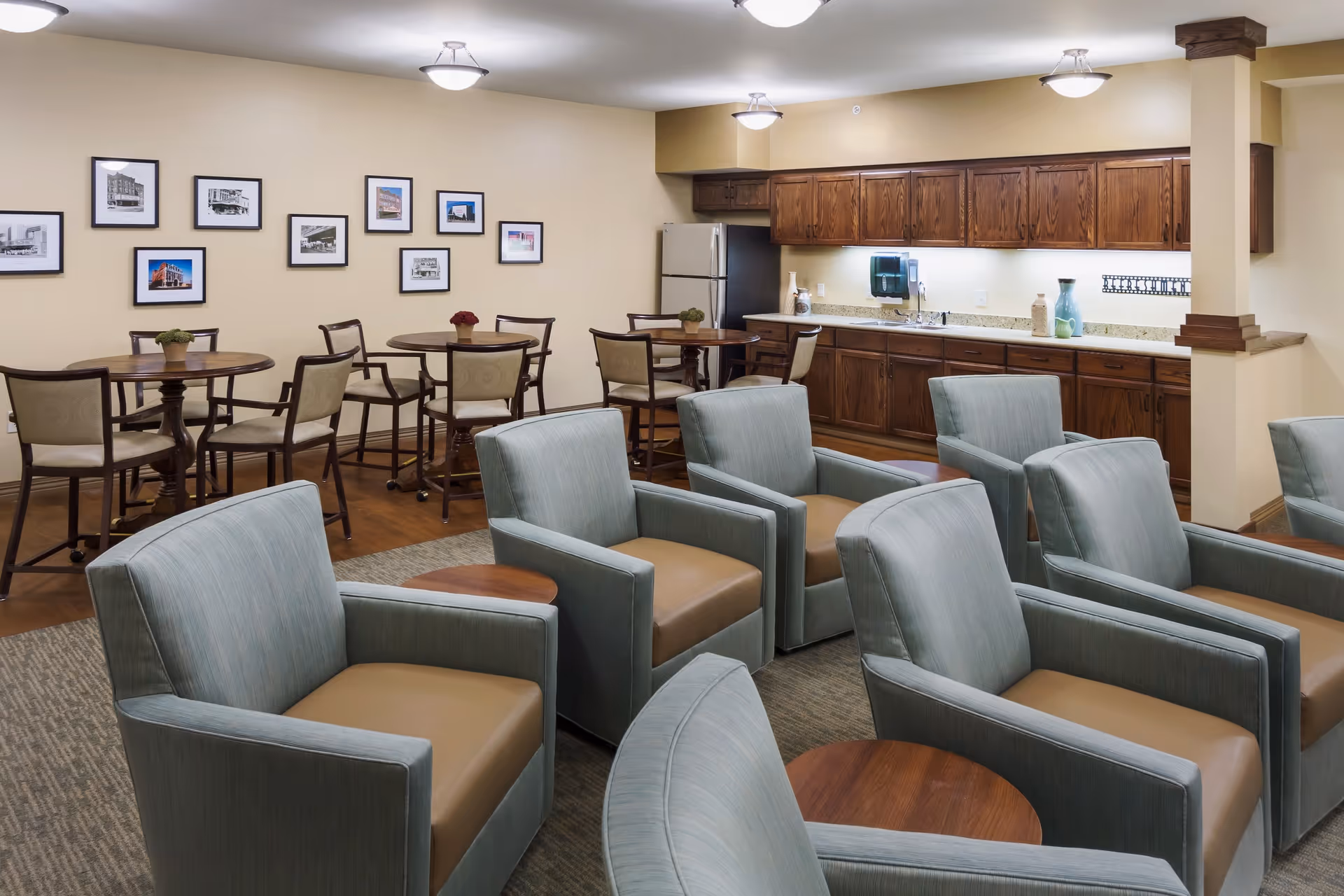 A common area in a senior living facility featuring several light blue armchairs with tan cushions arranged around small wooden tables. In the background, there are round wooden tables with chairs and a kitchenette with wooden cabinets, a refrigerator, and a countertop with decorative vases. The walls are decorated with framed black and white and color photographs.