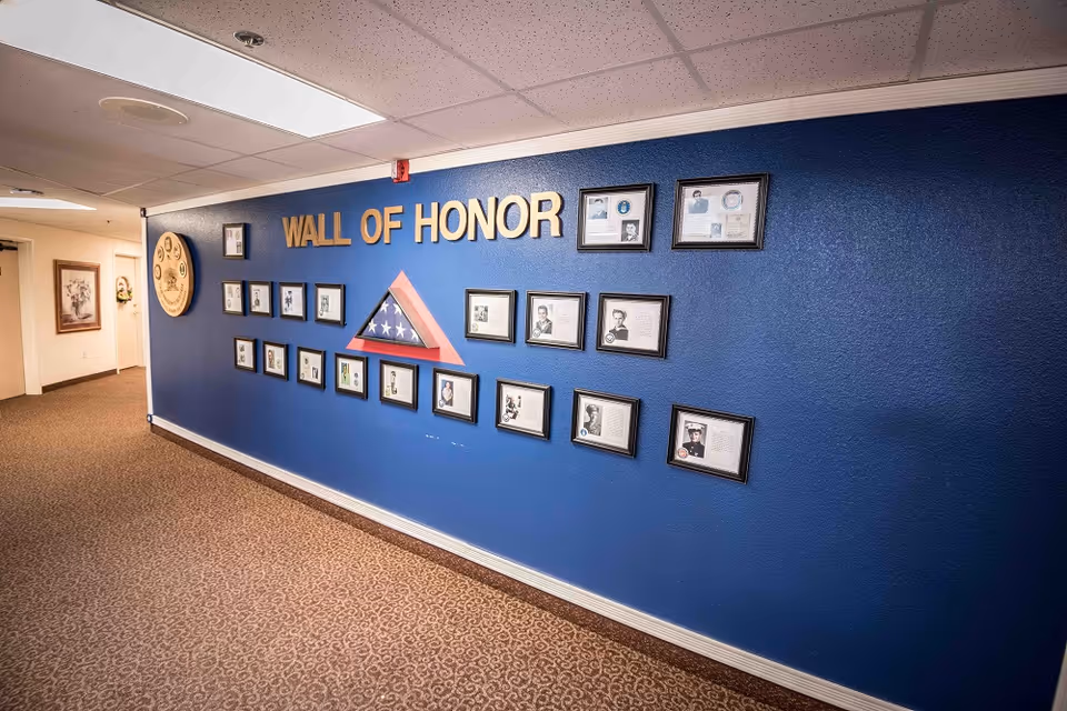 A hallway with a blue wall displaying a 'Wall of Honor' featuring framed photographs and documents, along with a folded American flag in a triangular case. The floor is carpeted with a patterned design, and the ceiling has recessed lighting panels.