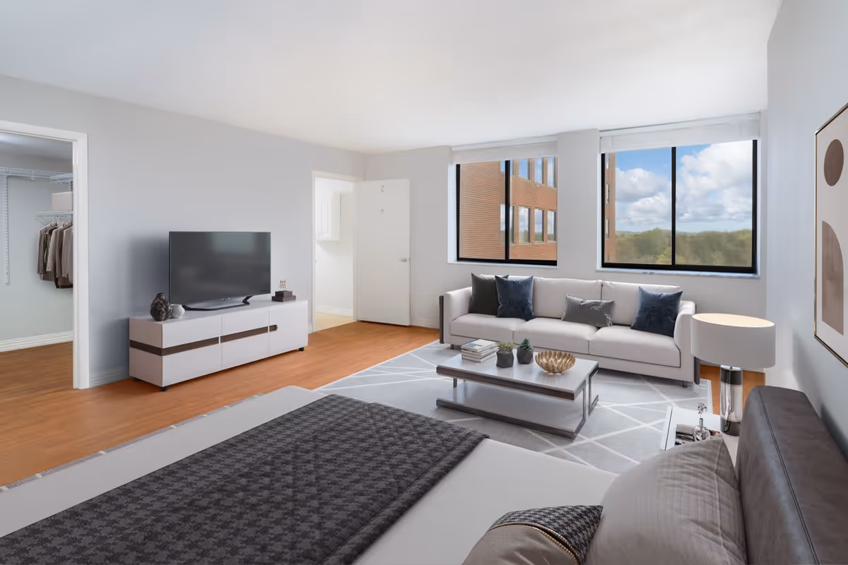 Bright studio apartment interior with a bed in the foreground, a sofa and coffee table by large windows, and a TV on a low console.