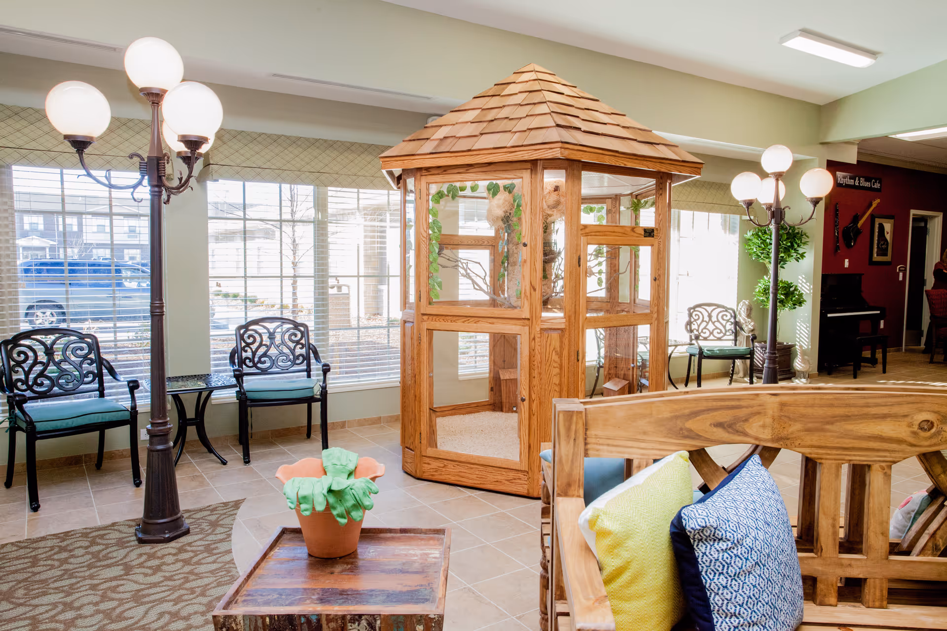 A bright and cozy common area in a senior living facility featuring a wooden gazebo-like structure with glass panels, decorative chairs with cushions, a wooden bench with colorful pillows, a small table with a potted plant, and vintage-style floor lamps. Large windows allow natural light to fill the room.