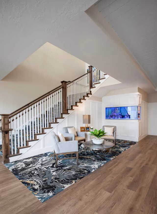 Bright foyer with a wooden staircase, a seating area of armchairs and a round coffee table on a patterned rug, and a built-in aquarium.
