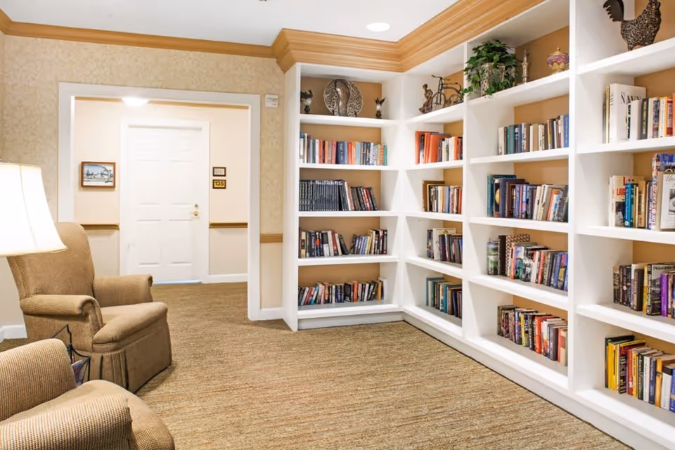 A cozy reading nook in a senior living facility featuring beige armchairs, a floor lamp, and white built-in bookshelves filled with books. The walls have a light patterned wallpaper and there is a white door with the room number 135 visible in the background.