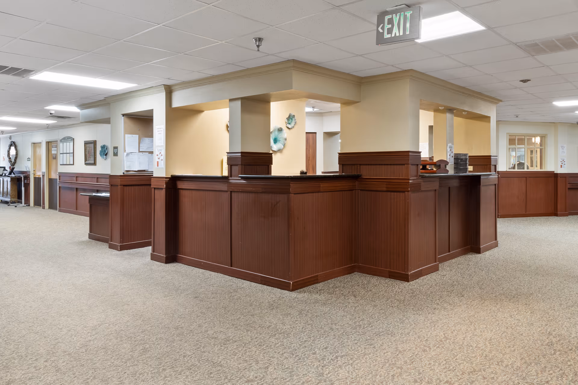 Interior view of a senior living facility reception or nurse station area with wood-paneled counters and beige walls. The space has carpeted floors, ceiling tiles with fluorescent lighting, and an illuminated exit sign above the station. Various wall decorations and notices are visible in the background.