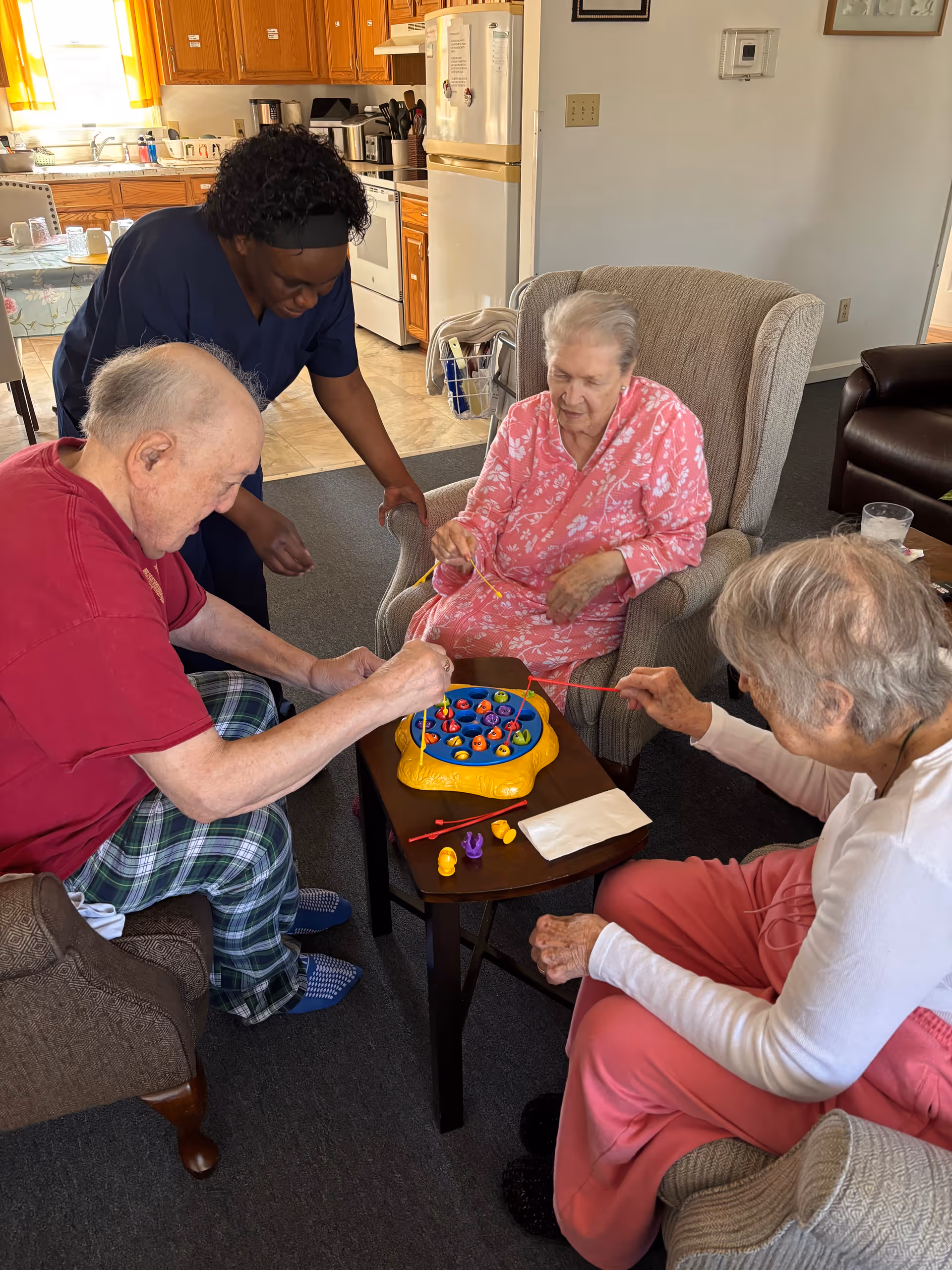 Three elderly residents sitting in a living room playing a tabletop fishing game while a caregiver leans in to assist.