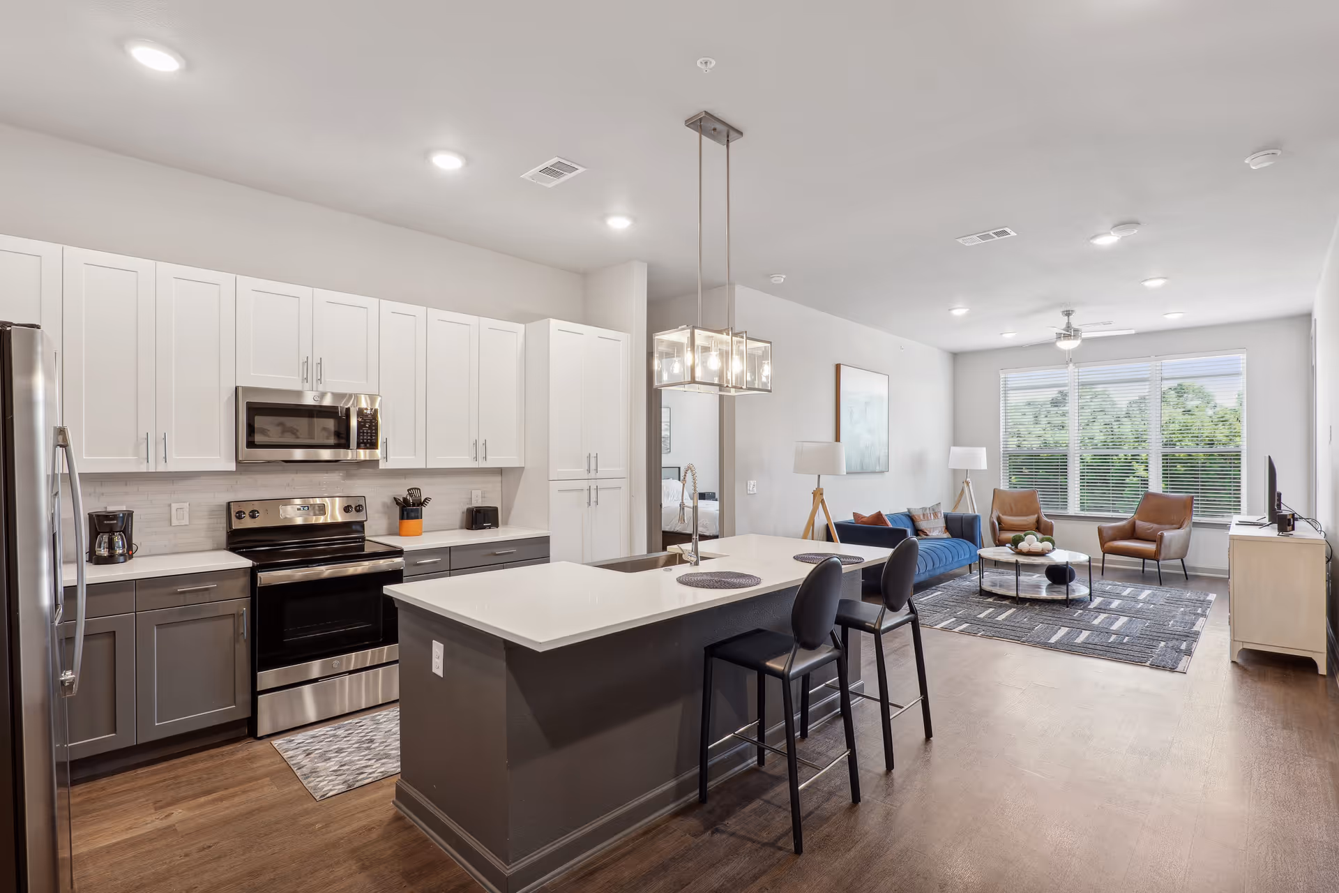 Modern open-concept kitchen and living room area with white and gray cabinetry, stainless steel appliances, a kitchen island with two black bar stools, and a living room furnished with a blue sofa, two brown armchairs, a round coffee table, and large windows letting in natural light.