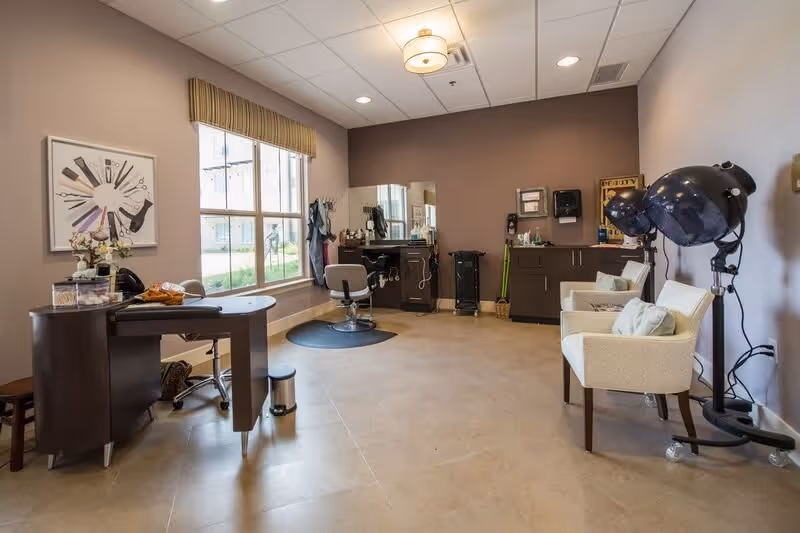 A well-lit salon room with a large window, a styling chair in front of a mirror, a desk with a chair, two white cushioned chairs with pillows, and two black hair dryers on stands. The walls are painted in neutral tones and the floor is tiled.