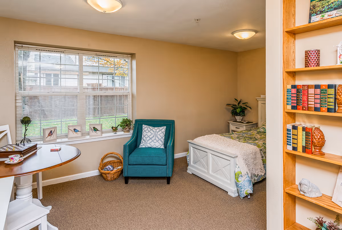 A cozy senior living bedroom with a large window featuring blinds and bird decorations on the windowsill. The room includes a teal armchair with a patterned cushion, a white wooden bed with a colorful bedspread and a white throw blanket, a small white nightstand with a plant, a round wooden table with a chessboard and teacup, and a wooden bookshelf filled with books and decorative items.