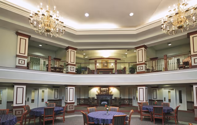 Two-story atrium-style common dining area with round tables, chandeliers, a fireplace, and balcony seating.