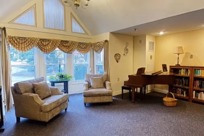 Bright lounge with two upholstered armchairs by large windows, a grand piano and a bookshelf with a lamp.