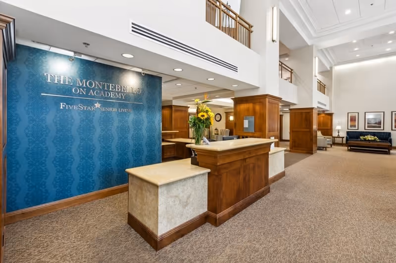 Reception area of The Montebello on Academy senior living facility featuring a wooden front desk with a flower vase, a blue accent wall with the facility's name and logo, and a spacious seating area with chairs and framed artwork in the background.