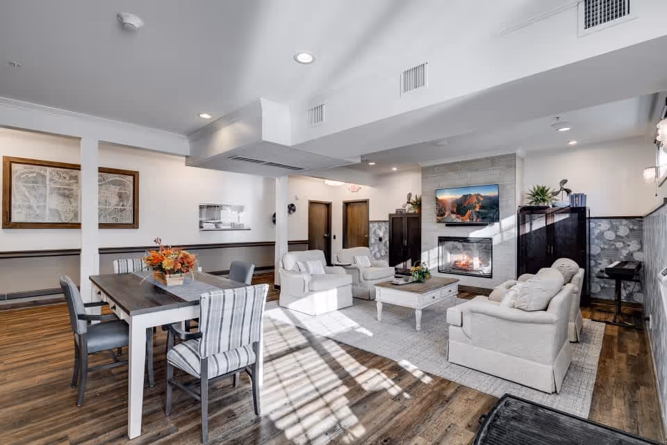 A bright and spacious senior living common area featuring a seating arrangement with two armchairs and a sofa around a coffee table, a fireplace with a mounted TV above it, and a dining table with six chairs. The room has wooden flooring, white walls, and decorative elements including plants and framed artwork.
