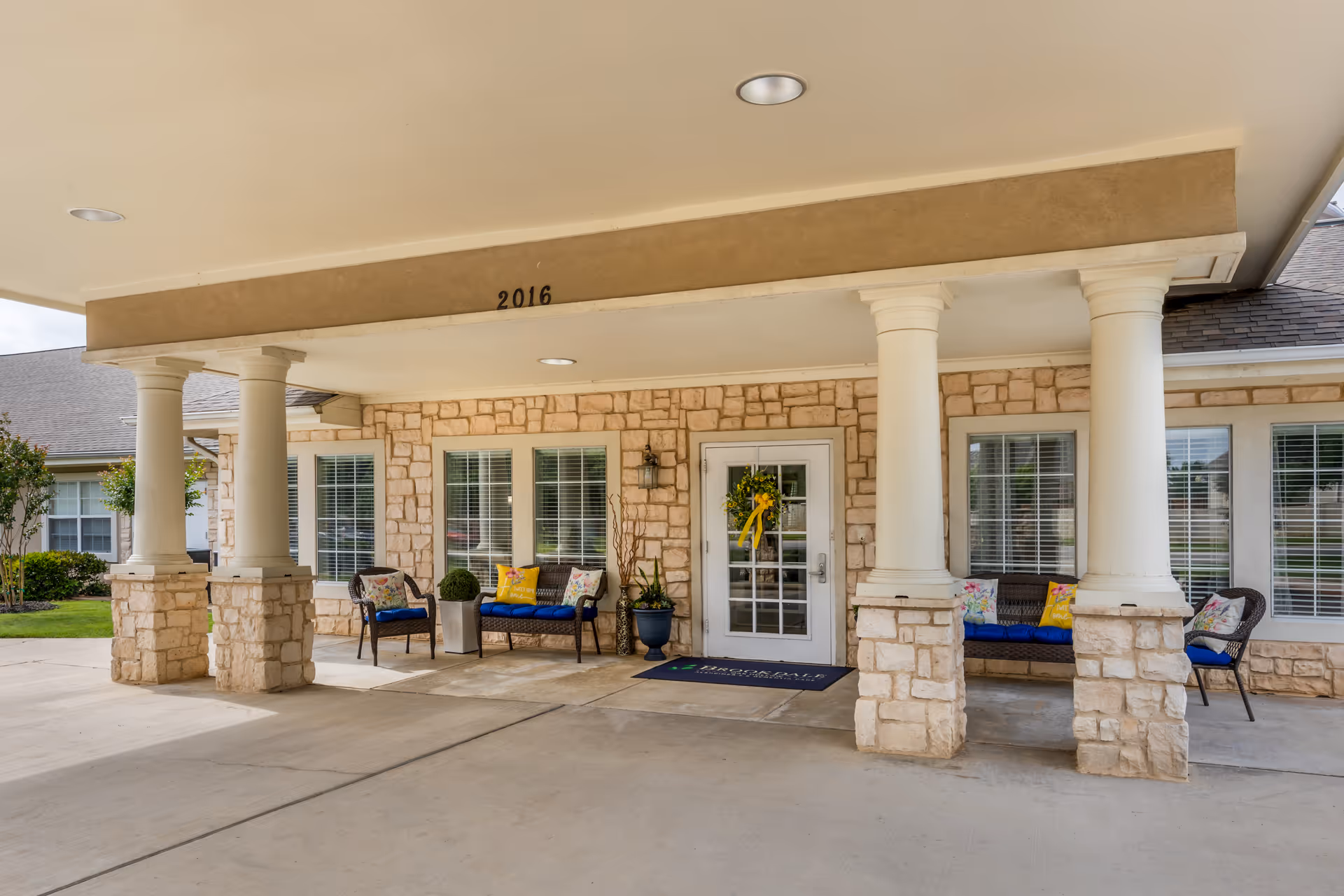 Covered front entrance of a stone-faced senior living facility with large white columns, outdoor seating, and a glass door decorated with a wreath.
