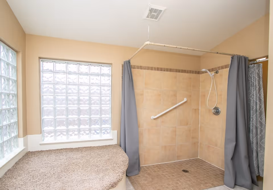 A bathroom with a walk-in tiled shower featuring a grab bar and a handheld showerhead. The shower area is enclosed by a ceiling-mounted curtain rod with gray curtains. Next to the shower is a built-in bench covered with beige carpet. The walls are painted beige, and there are large glass block windows allowing natural light into the space.
