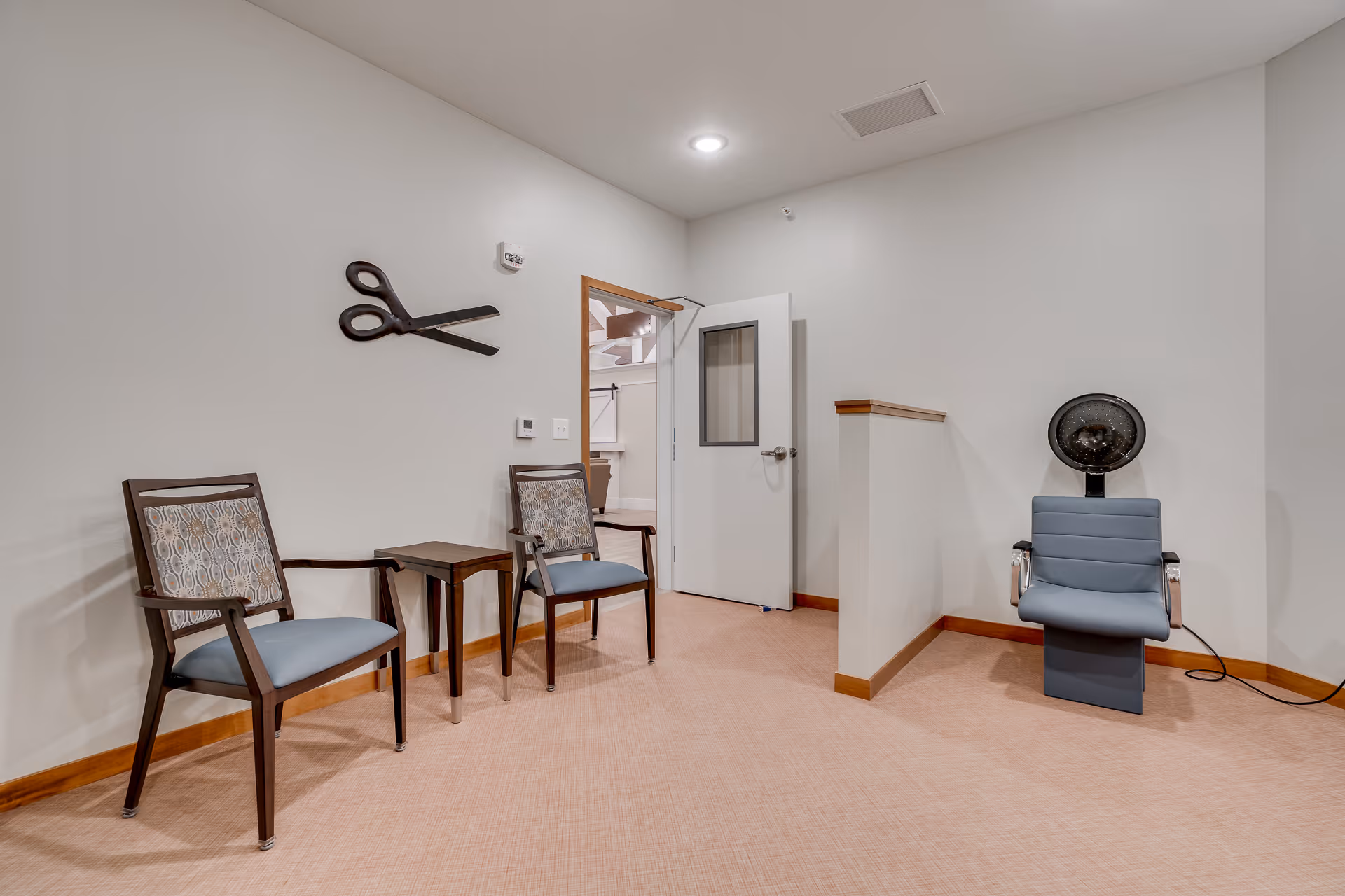 A small room with two wooden chairs with patterned cushions and a small wooden table between them on the left side. On the right side, there is a blue salon hair dryer chair. The walls are light-colored with a large decorative pair of scissors mounted on the left wall. A white door with a window is open, showing a glimpse of another room.