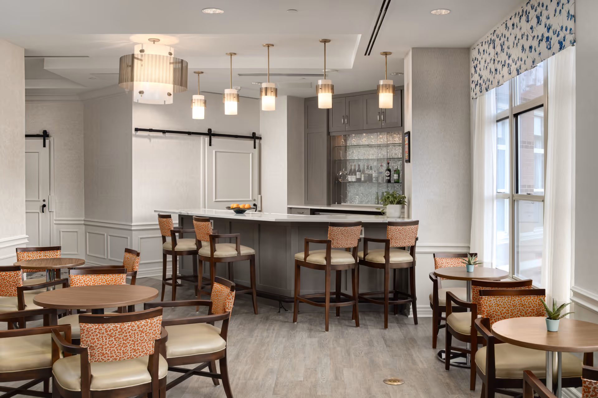 A bright and modern dining area featuring several round wooden tables with cushioned chairs that have patterned backs. There is a bar counter with high chairs in the background, pendant lights hanging from the ceiling, and large windows with floral valances letting in natural light.
