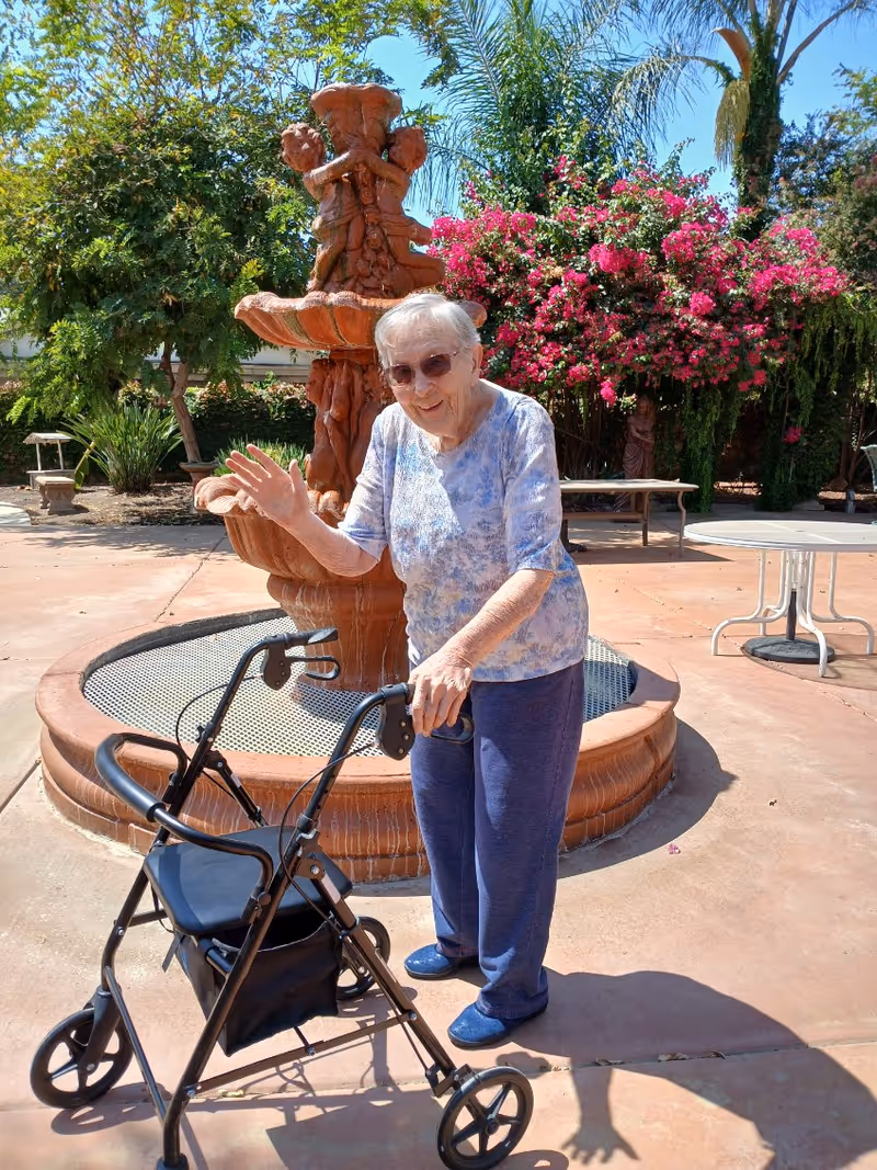 An elderly woman wearing sunglasses, a light blue patterned shirt, and blue pants is standing outdoors next to a black walker. She is smiling and waving with one hand. Behind her is a decorative fountain with sculpted figures, lush green trees, and vibrant pink flowers. There are also benches and a round table in the background.