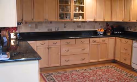 L-shaped kitchen with light wood cabinets, dark countertops, and a decorative rug.