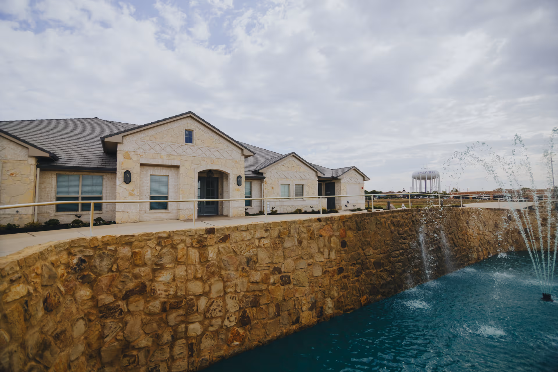 Stone-faced single-story building with a stone retaining wall and fountain pool in front under a cloudy sky.