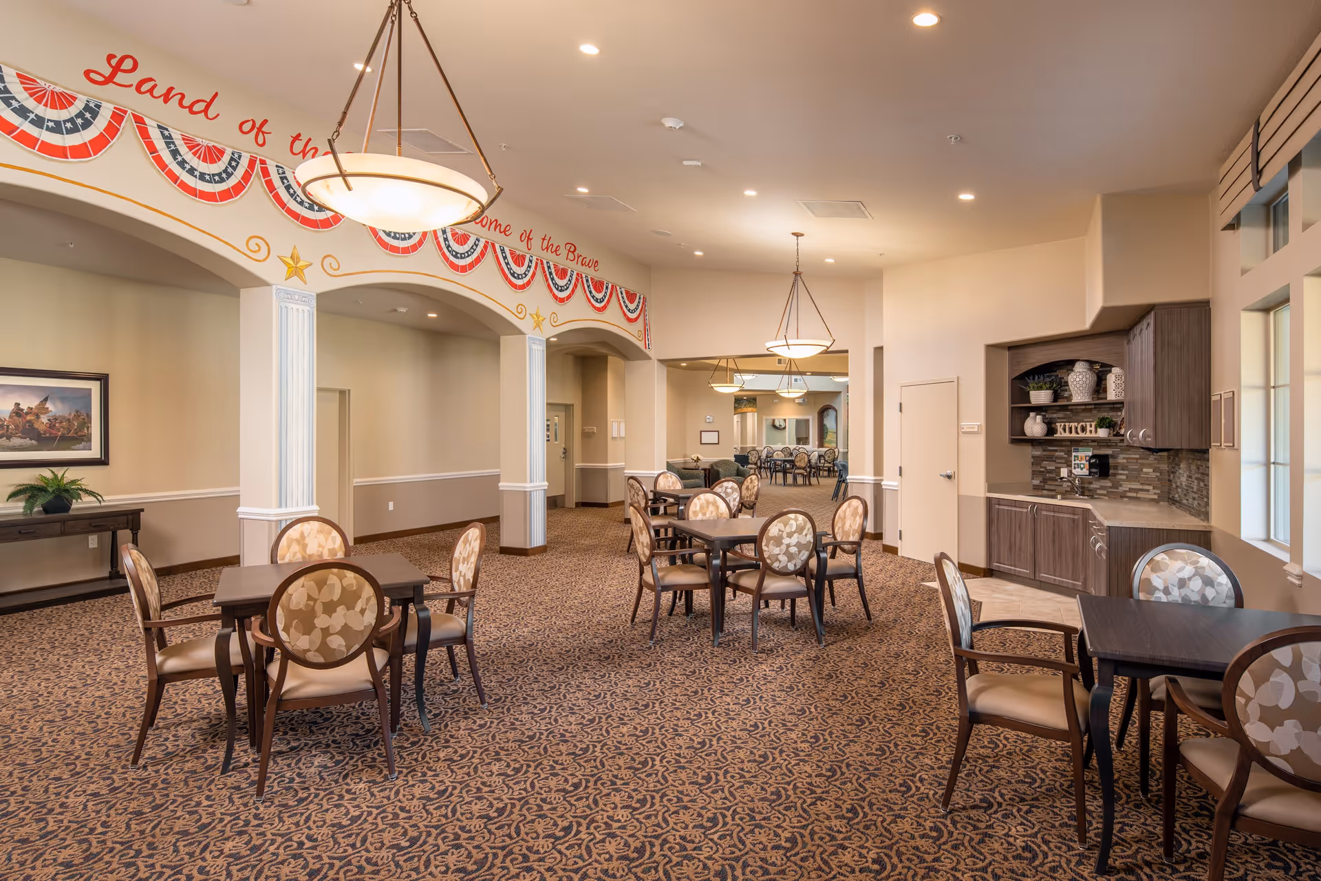 A spacious dining area in a senior living facility with multiple tables and chairs arranged on a patterned carpet. The room features warm lighting from hanging fixtures, a small kitchenette with cabinets and a sink on the right, and patriotic decorations with the phrase 'Land of the Free, Home of the Brave' above an archway. There is a framed painting and a plant on a console table against the left wall.