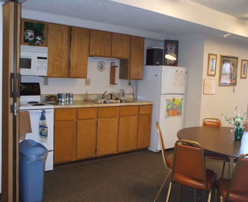 A small kitchen area with wooden cabinets, a white microwave, stove, and refrigerator. There is a double sink with a bottle of dish soap on the counter. A round dining table with four brown chairs is visible to the right, with a small vase of flowers on the table. The walls have framed pictures and a calendar.