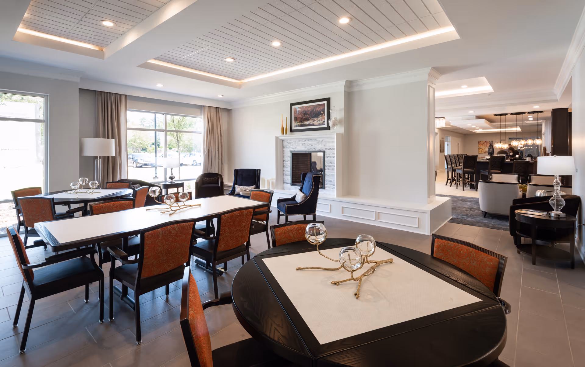 A bright and spacious common area in a senior living facility featuring multiple tables with chairs, decorative centerpieces, large windows with beige curtains allowing natural light, a white fireplace with a framed picture above it, and a seating area with armchairs. The room has a modern ceiling with recessed lighting and a view into an adjacent dining or lounge area.