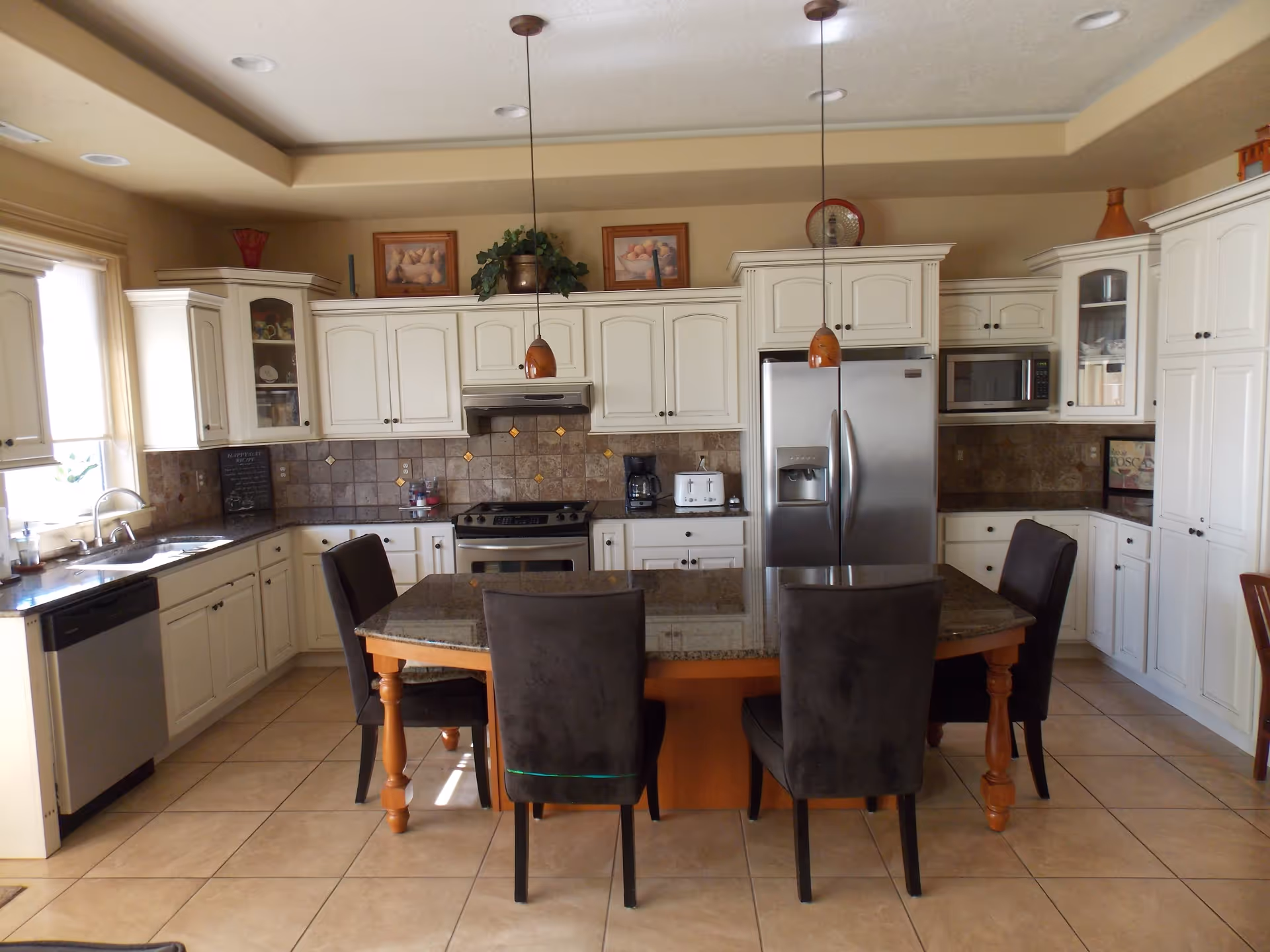 A spacious kitchen with white cabinetry, a stainless steel refrigerator, stove, microwave, and dishwasher. There is a large island with a granite countertop surrounded by four dark upholstered chairs. The kitchen has tiled floors and a beige tiled backsplash, with two pendant lights hanging above the island. Decorative items and framed pictures are placed on top of the cabinets.