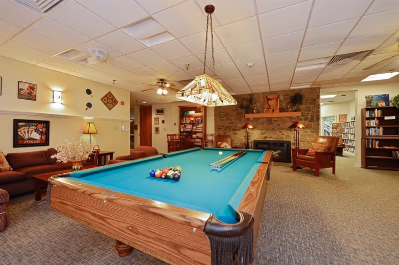 Game room with a pool table, sofas, armchairs, a stone fireplace and bookshelves.