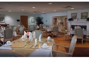 A spacious dining room with multiple tables set for a meal, featuring white tablecloths, folded napkins, cups, and silverware. The room has wooden flooring, upholstered chairs, framed artwork on the walls, and a fireplace in the background.